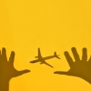 Photograph of a shadow of a plane and two tough guys reaching for the plane, against a yellow wall.