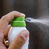 Close-up photograph showing a hand pressing the button on a can of mosquito repellent spray. The spray catches the light when it comes out of the can.