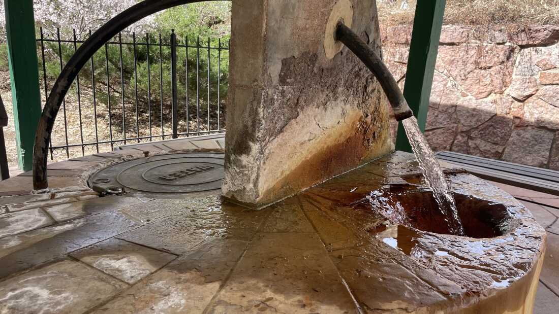An arcing metal pipe lets water flow into a stone basin. Reddish-brown rust, the product of oxidization, covers both the basin and the mouth of the pipe.