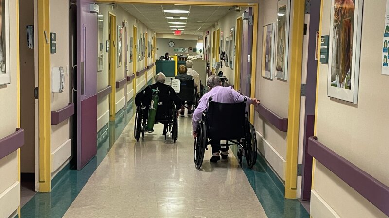 Three people in wheelchairs navigate the hallway of a nursing home. 