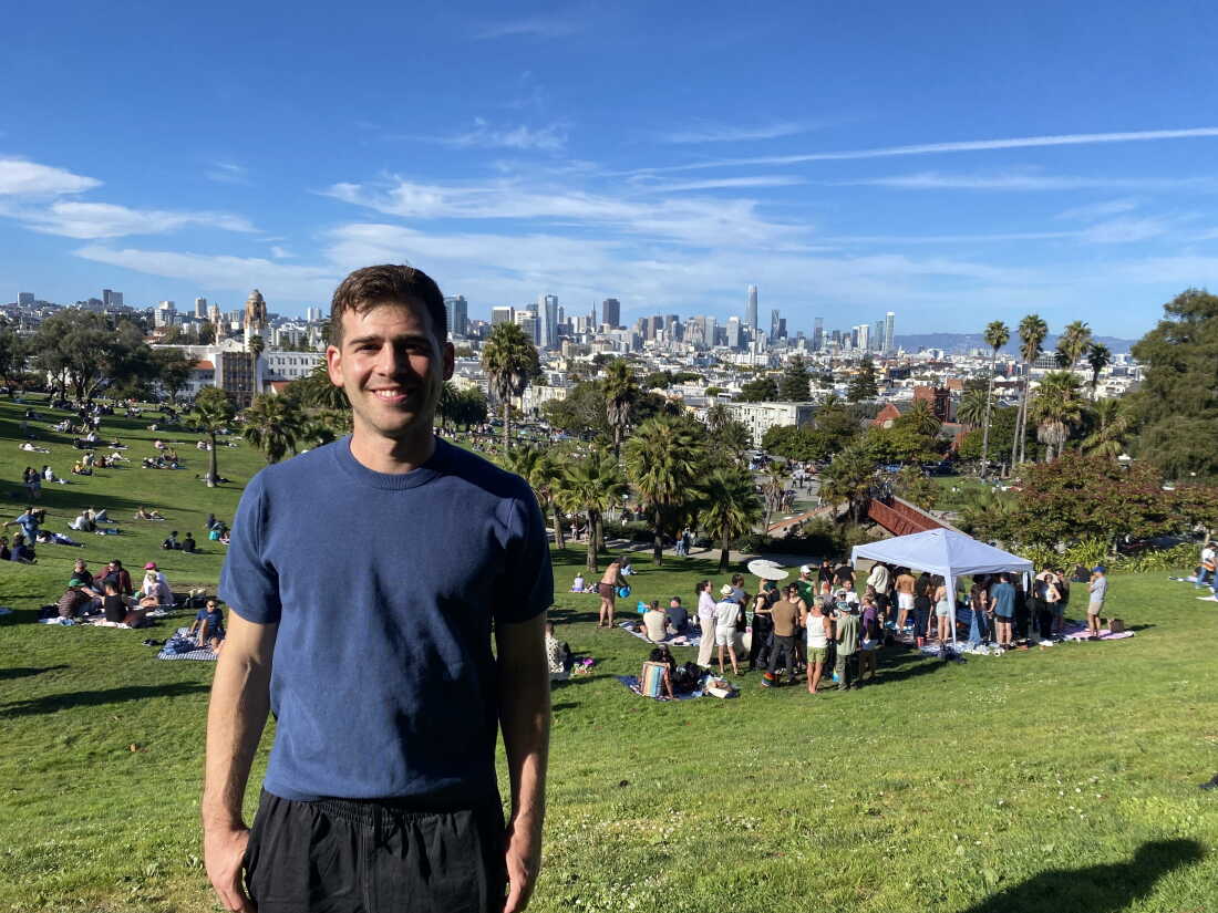 Alex Brenner stands on a hillside at Mission Dolores Park. A vista view of San Francisco is in the background.