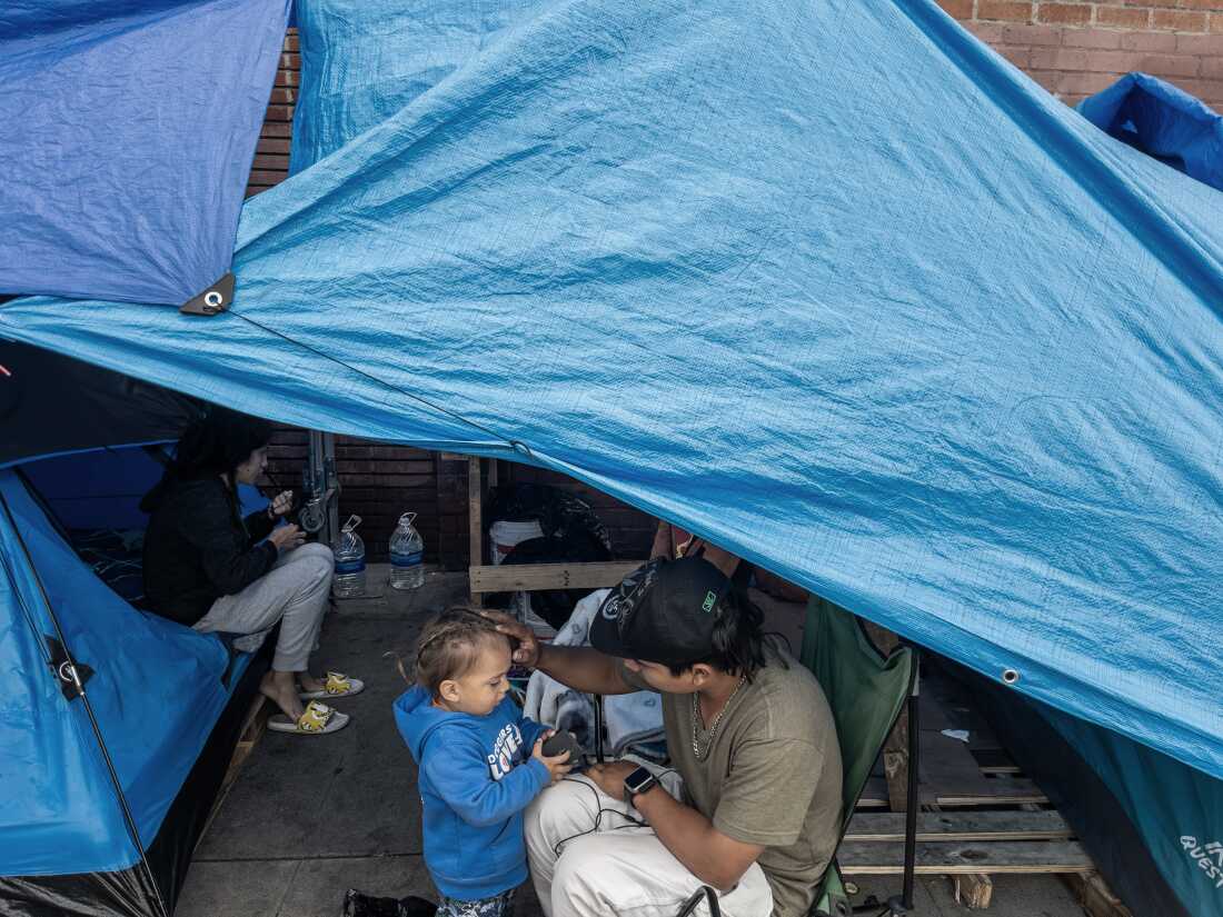A small child with braided light brown hair wearing a blue hoodie interacts with a man in a baseball hat and brown t-shirt. They are under a blue tarp rigged to give them some shelter. A woman is in the background sitting near some large bottles of water.