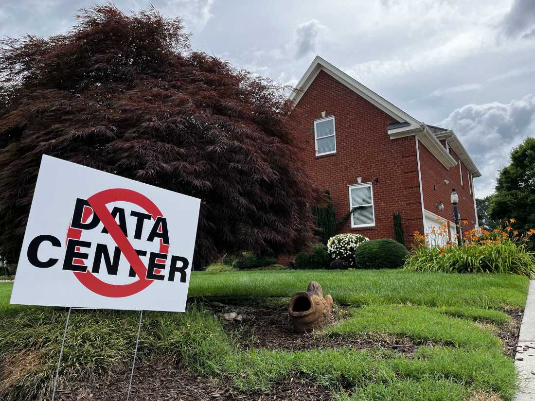 This photo shows a white yard sign that says in all caps, "NO DATA CENTER!" A red ring with a slash through it appears on top of the text. Stretching behind the sign is a grassy green yard, and in the background stands a brick house.