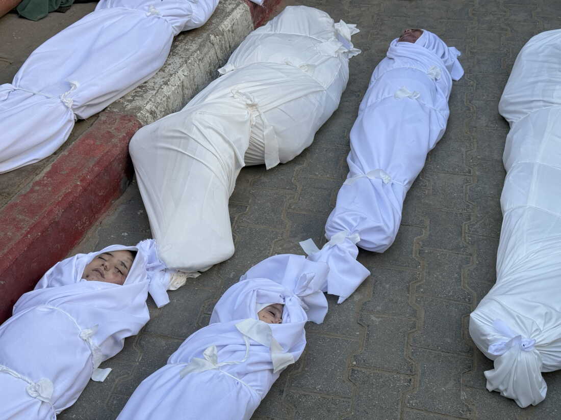 The bodies of six people wrapped in white shrouds lie on the ground in two rows at a morgue at Al-Shifa Hospital in Gaza City on November 20, 2025. At least two of the bodies appear to be young children, with their faces uncovered and eyes closed.