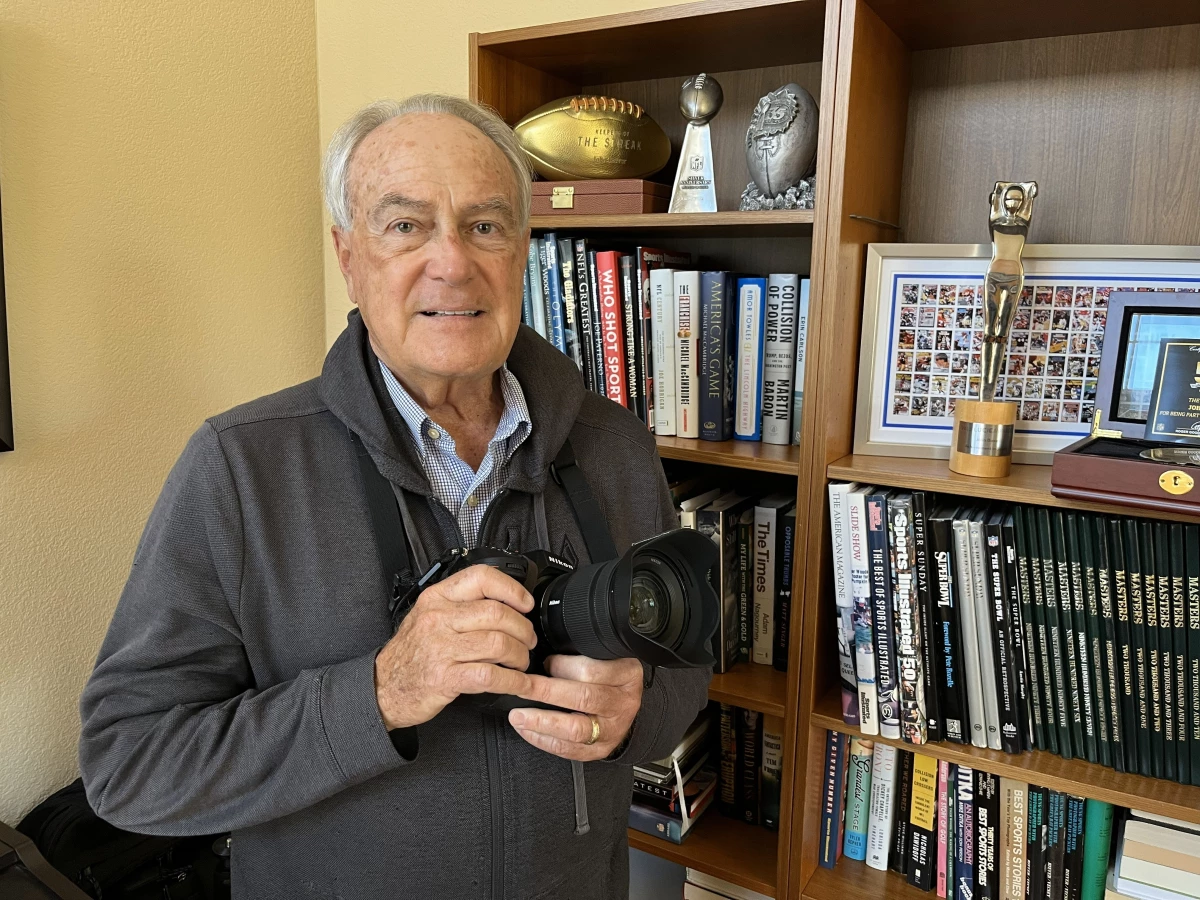 John Biever poses with a camera in front of some memorabilia at his San Diego home. He covered his first Super Bowl at the age of 15 and, on the verge of turning 75, he says Super Bowl LX will probably his last.