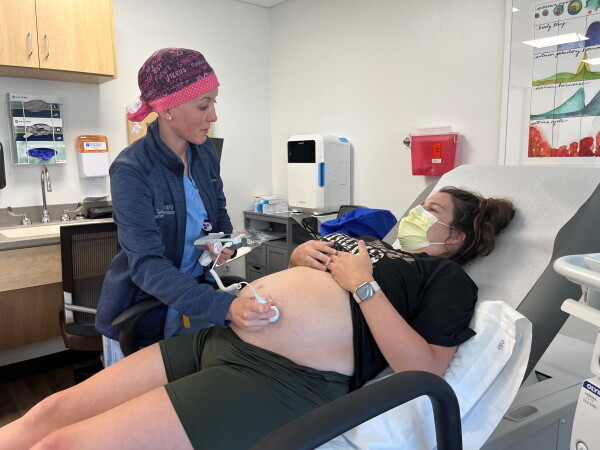 Addie Comegys, a pregnant woman, lies on a medical exam table with her T-shirt partially lifted while obstetrician-gynecologist Taylar Swartz presses an ultrasound scanner on Comegys' exposed abdomen.