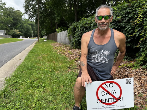In this photo, Lee DaMore is kneeling on the grass behind a white yard sign that says in all caps, "NO DATA CENTER!" A red ring with a slash through it appears on top of the text. DaMore is wearing a gray tank top, shorts and sunglasses.
