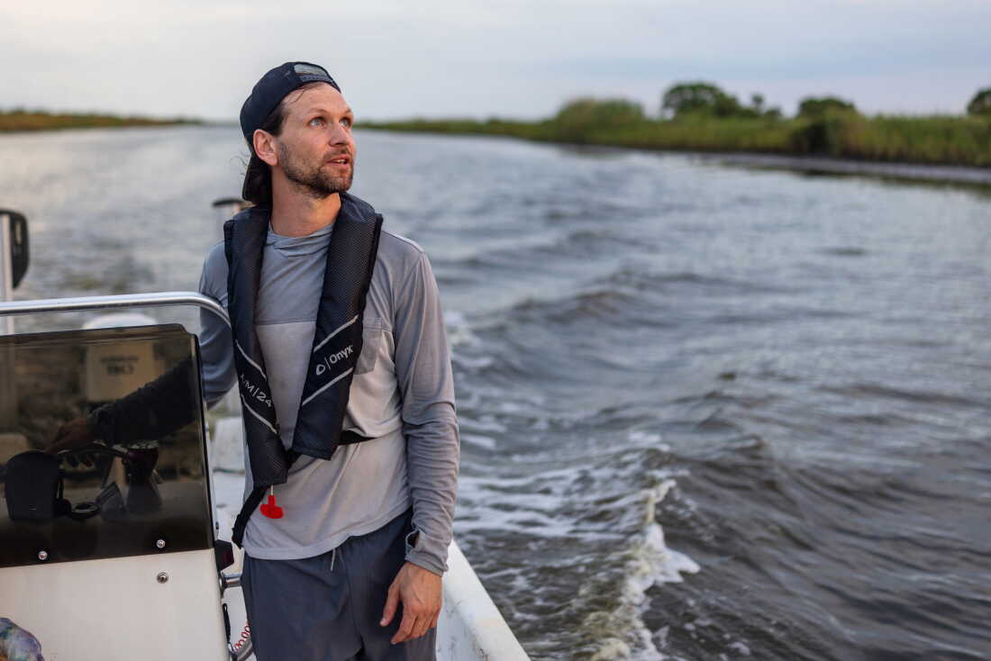 A man in a long-sleeved shirt and baseball cap on backwards stands on a small boat and behind him is a long stretch of water and green brush on the shore.