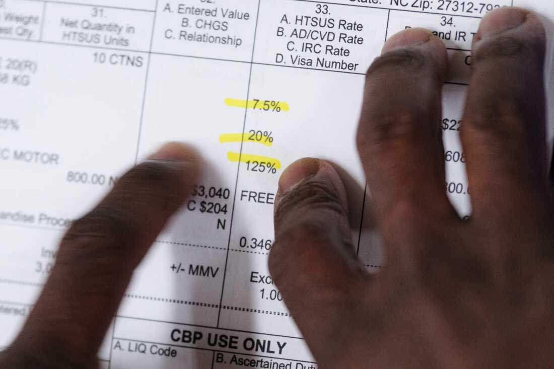 Joshua Esnard's fingers point to the tariff line items on a paper tariff document. Three percentages are highlighted in yellow — 125%, 20% and 7.5% — summing to 152.5%.
