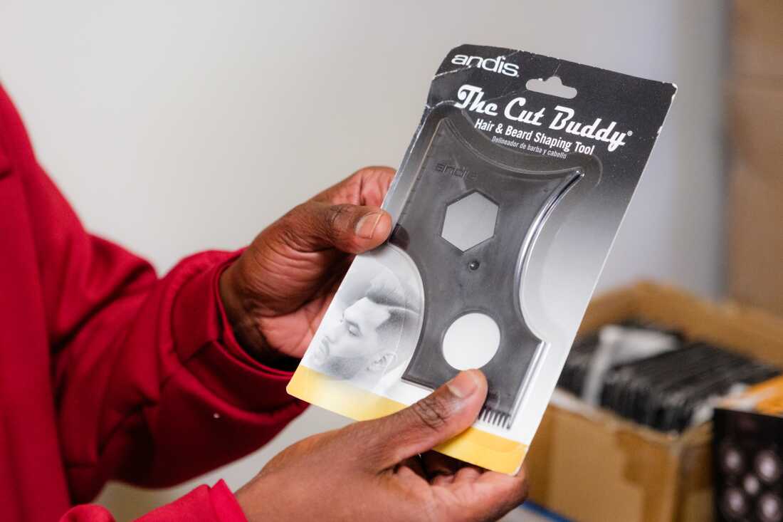 Joshua Esnard's hands hold The Cut Buddy hair and beard-shaping tool in its packaging. The tool is a flat, black plastic stencil with curves, points and cutout shapes.