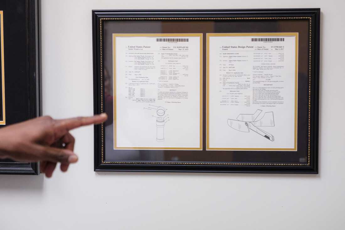 A certificate for a patent that Joshua Esnard created hangs in a dark frame on the wall of his office inside The Cut Buddy warehouse. On the left side of the frame, a finger points to the framed certificate.