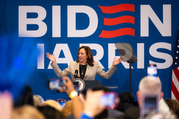 A wide shot of Vice President Kamala Harris at a campaign rally at a high school on July 18. She has both of her arm raised and outstretched as she addressed a crowd in front of a large "Biden-Harris" poster.