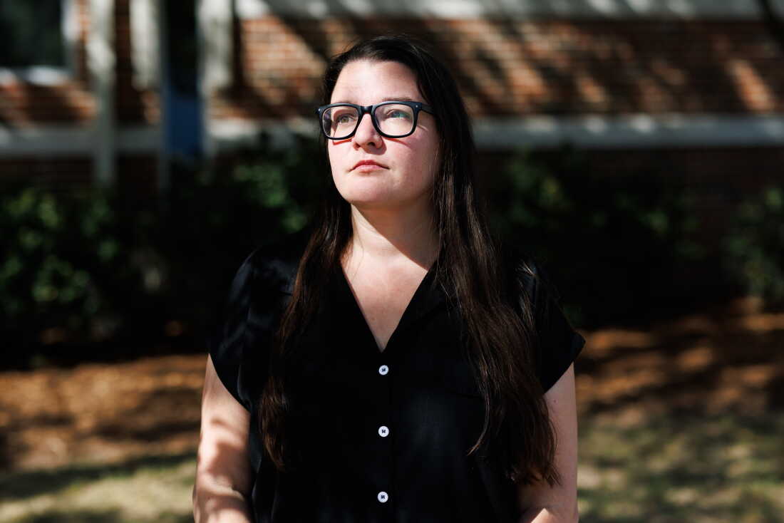 Leigh Mazur stands outside, with a portion of a blurry brick building in the background. Photographed from about the waist up, she's wearing glasses and a black, short-sleeved outfit.