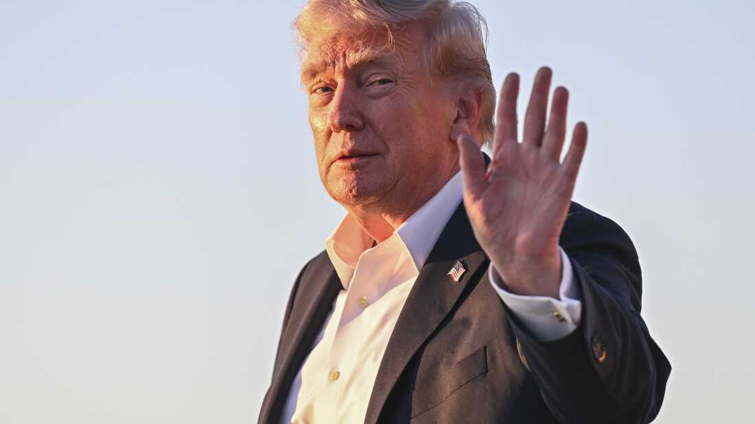 President Trump waves while wearing a black suit and a white button-up shirt. He is seen from the chest up.