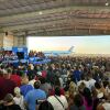 Cette photo montre une scène d'un rallye de Kamala Harris et Tim Walz à Détroit le 7 août. Beaucoup de gens sont encombrés ensemble dans un hangar d'avion, face à une scène où Harris parle à un pupitre. Le hangar est ouvert à l'extérieur, et à l'extérieur se trouve Air Force Two et un ciel bleu.