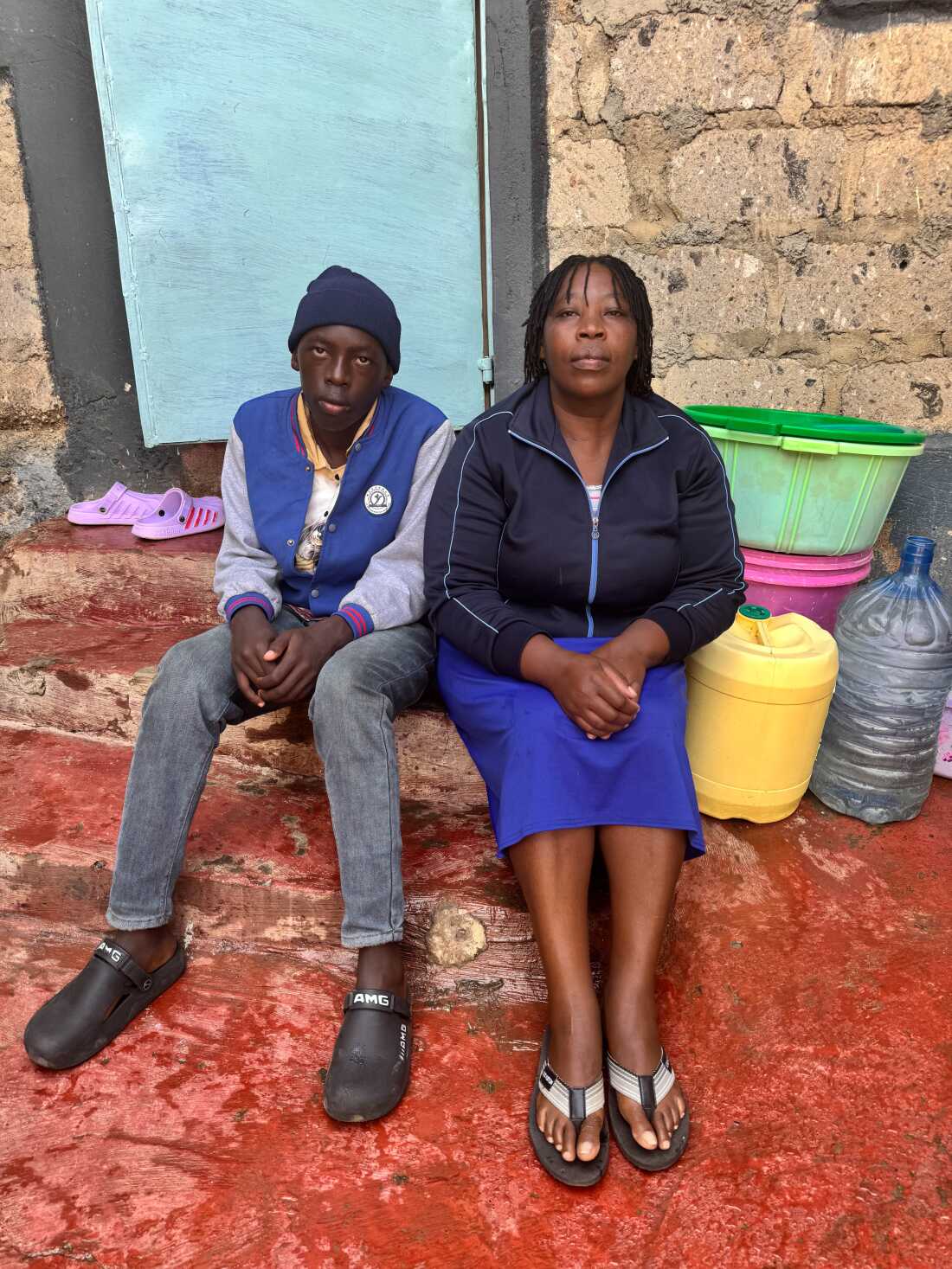 Purity’s younger brother, James Gitau Mwai, and her mother sit on steps outside their home in Nairobi, Kenya. Behind them are plastic basins and large plastic containers for holding water.