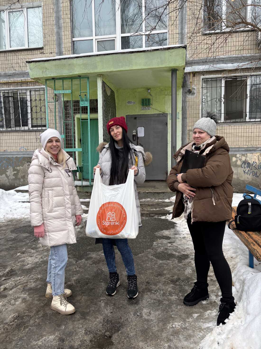 Three women who are volunteers with the aid group Starenki stand outside the entrance to a multistory apartment building. They are wearing winter coats and knitted caps. The woman in the middle is holding a large plastic bag that says Starenki on it.