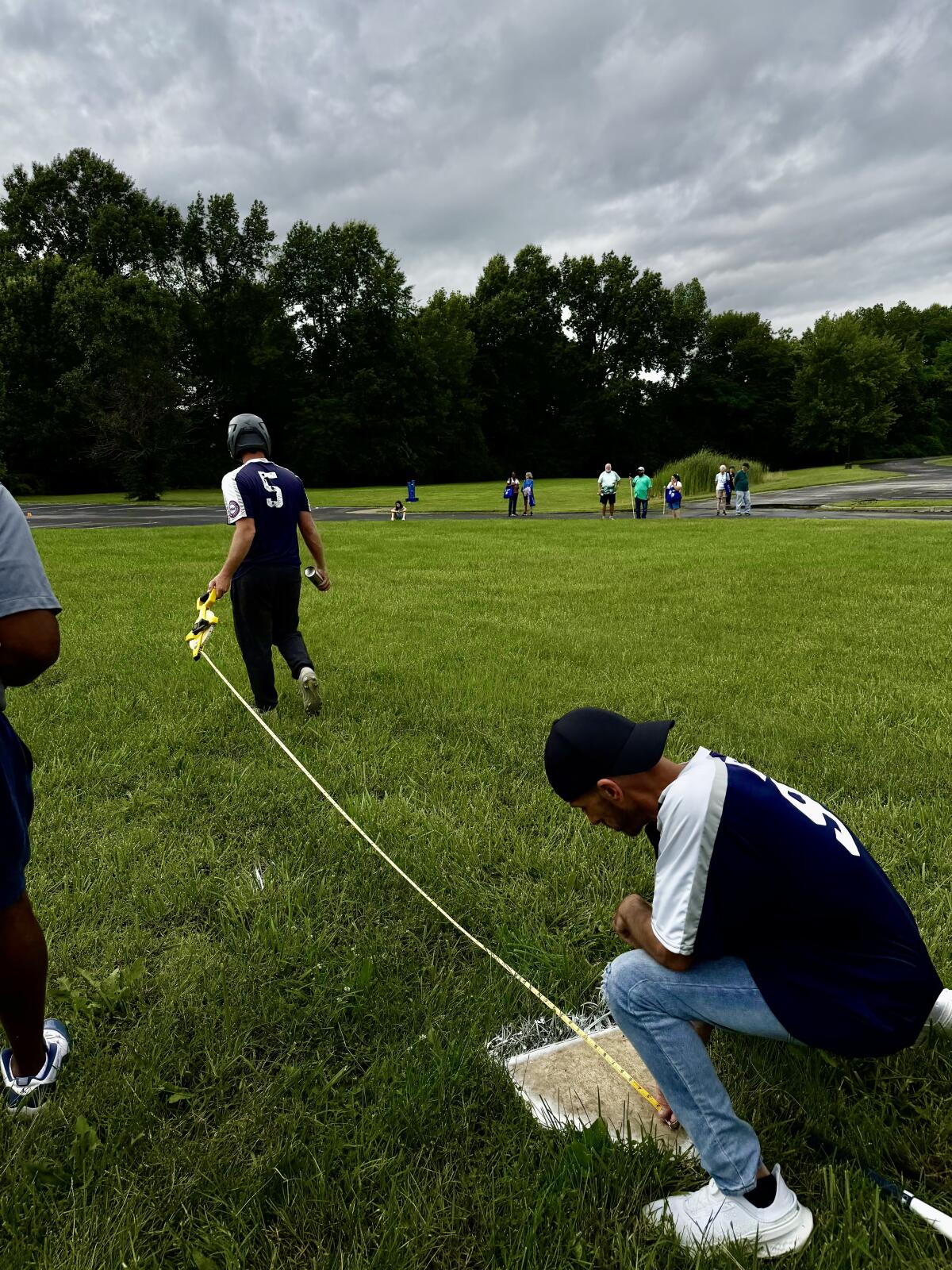 Jared Woodard and another player measure out the distance from home plate to first base with a handheld field-measuring tape. The skies are overcast and the grass is wet.