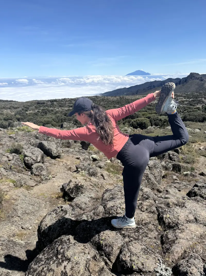 Sophie Rottenberg is shown with a rugged mountain vista behind her. She is balanced on one leg in yoga's "dancer" pose.