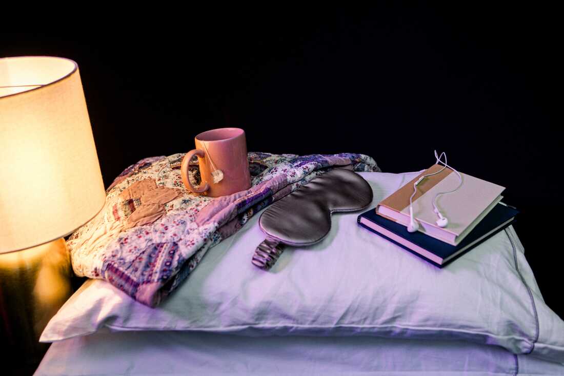 A pink mug of hot tea, a gray silk sleep eye mask, two books and some wired headphones are laid out on top of two stacked pillows next to a table lamp that's turned on, emitting warm light. All the objects are photographed in a studio against a black backdrop.