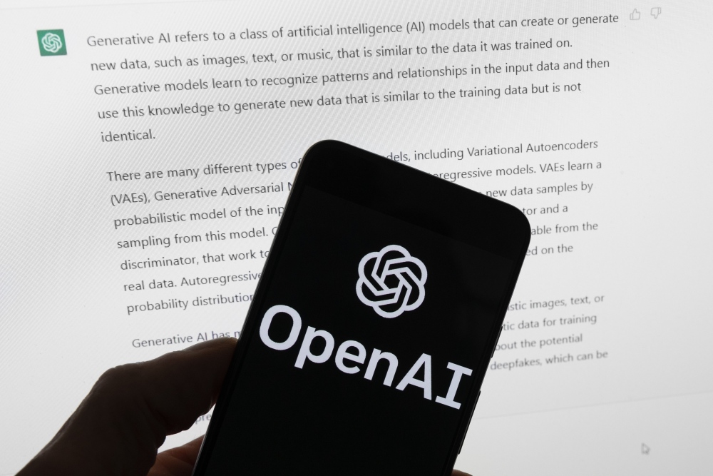 OpenAI's logo is on the screen of a mobile phone that's being held in front of a computer screen displaying output from ChatGPT. (AP)