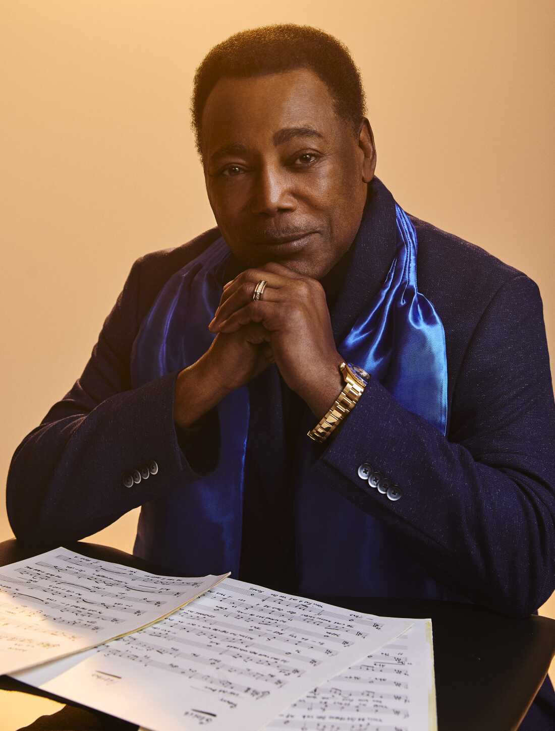 George Benson sits at a table with his sheet music in front of him. He is wearing a dark blue blazer jacket with a light blue satin scarf.