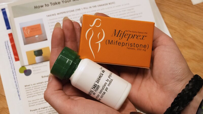 A person holds two packages of medicine in their hands, one in an orange box, the other in a white plastic pill bottle. In the background is an instruction sheet, with the headline "How to take your medication" partially visible.