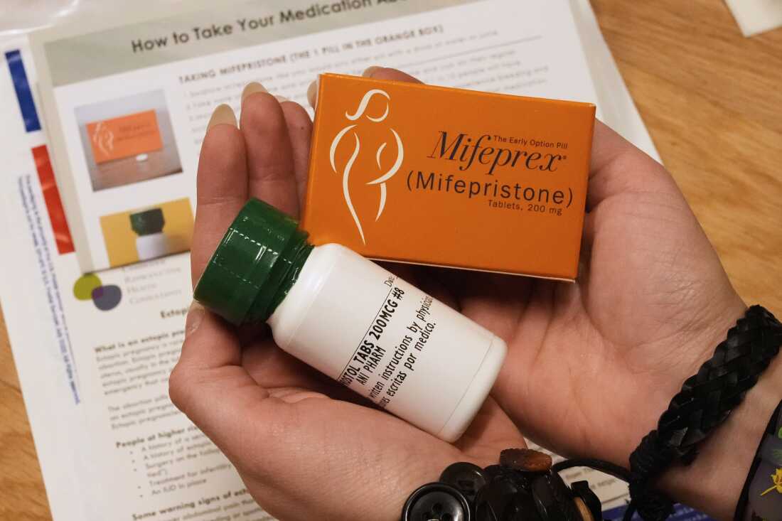 A person holds two packages of medicine in their hands, one in an orange box, the other in a white plastic pill bottle. In the background is an instruction sheet, with the headline "How to take your medication" partially visible.