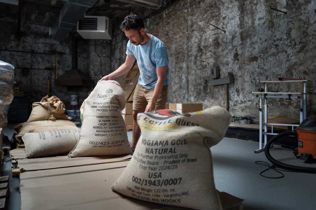In this photo, Jeff Yerxa stands in a warehouse with several large bags of coffee beans in burlap-style sacks. A sack in the foreground says "Product of Brazil" on it, and other sack says "Sancoffee Brasil" on it.