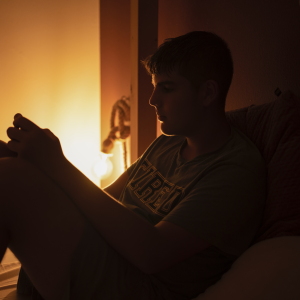 A teen boy is shown sitting up in his bed, looking at his phone. The room is dark except for dim lamplight.