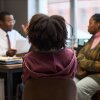 Black man appearing middle aged sitting at desk talking to Black woman and Black child in office setting, principal discussing school matters with parent and student.