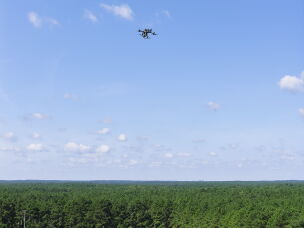 An aerial view of an Ocean County Sheriff's Office DJI Matrice 300 RTK drone with a thermal-capable camera payload at the Ocean County Fairgrounds with the New Jersey Pinelands stretching into the distance. A propeller plane can be seen below and right of the drone.