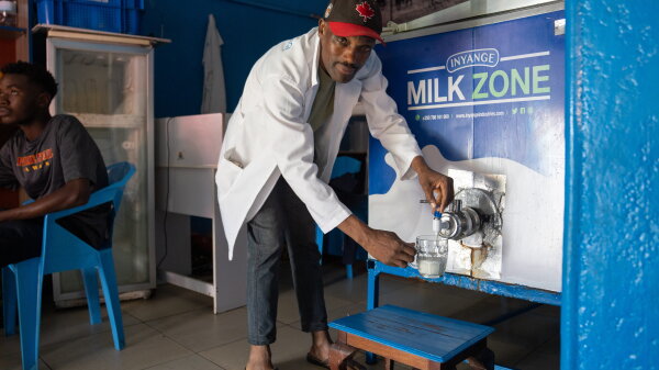 A man wearing a white long jacket, dark-colored pants and a baseball cap pours milk from a tap into a cup. 