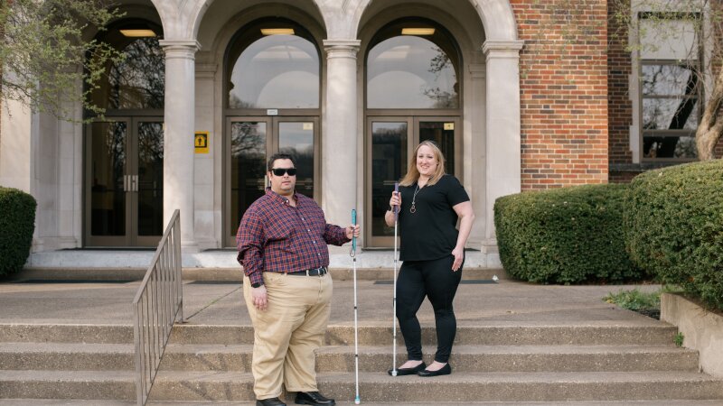 ALT TEXT: A 34 year old white man with short, black hair, dark glasses, and a plaid shirt stands to the left. A 43 year old woman with shoulder length blonde hair and an all black outfit, stands to the right. They’re both holding canes on stairs in front of a brick building.