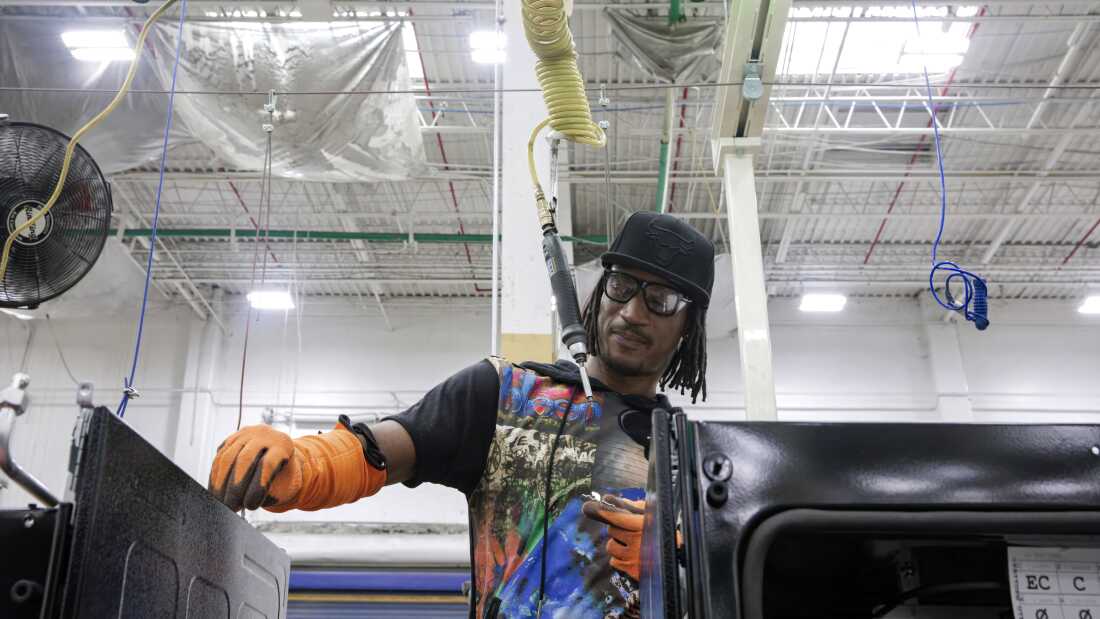 Kwame Crockett is a Black man and he is wearing a black baseball cap, safety glasses and a colorful shirt as he assembles parts of an oven.
