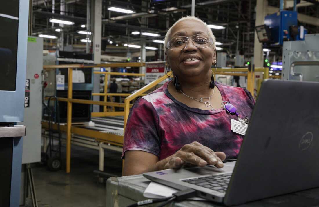 Ruth Ransom, 68, is a woman with short white hair. She is wearing safety glasses and standing behind a laptop on the factory floor.