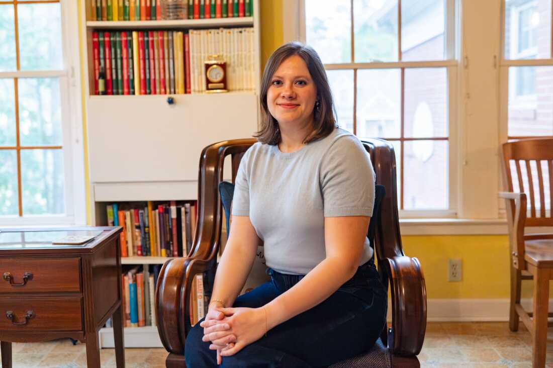 Caroline sits in a vintage wooden chair with windows and a bookcase behind her.