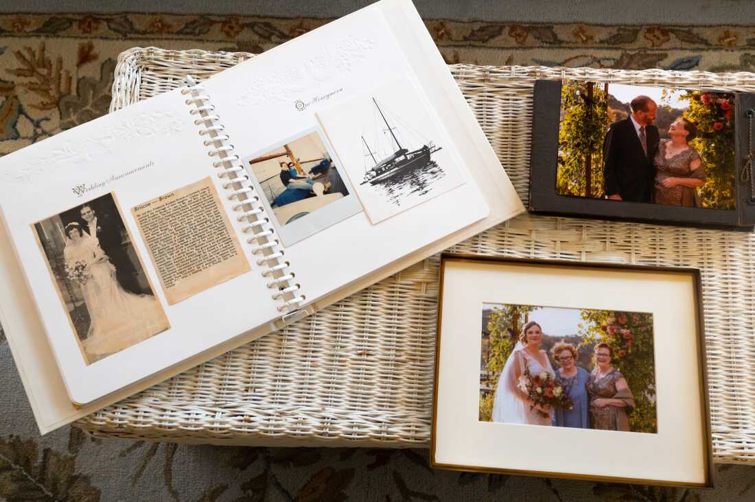 A white photo album has a black and white photo from Barbara's wedding along with a newspaper clipping. There is a framed photo of Caroline at her wedding with her grandmother and mother. And a photo of Cynthia looking at her husband at Caroline's wedding. The photos are displayed on a white wicker table.