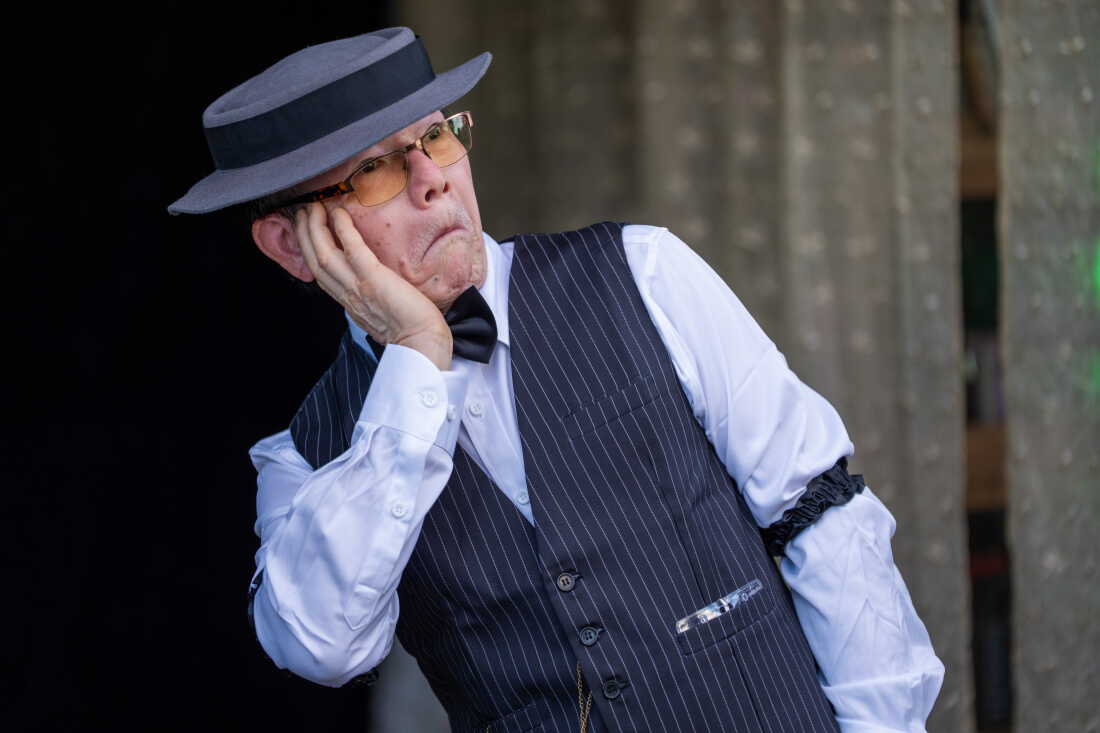 Khoi Nguyen, dressed as Buster Keaton in pinstripe vest over a button down shirt, a porkpie hat and a bowtie, puts his hand on his face.