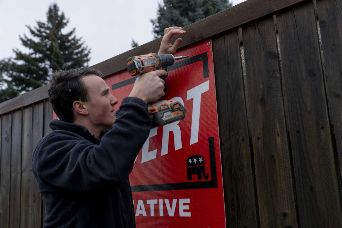 Montana state Rep. Lukas Schubert uses a drill to hang a campaign sign on a wooden fence in Kalispell.
