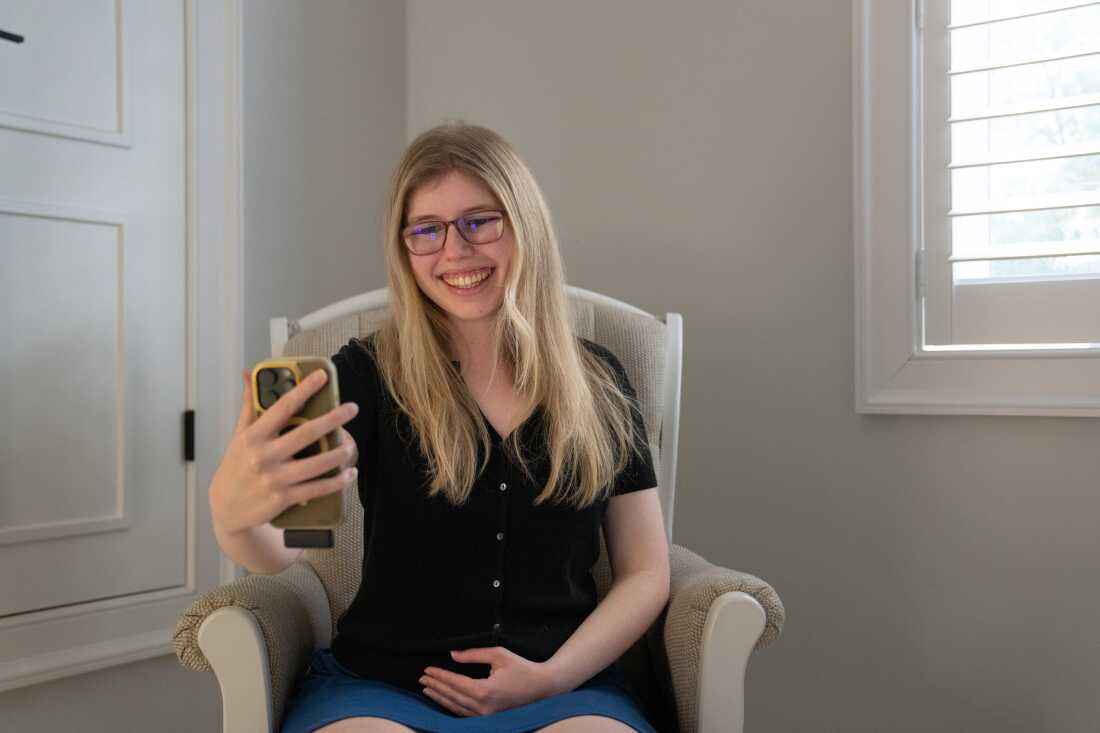 Kaleigh Brendle sits in a beige rocking chair while holding a smartphone in front of her body. She's looking at the phone and smiling.