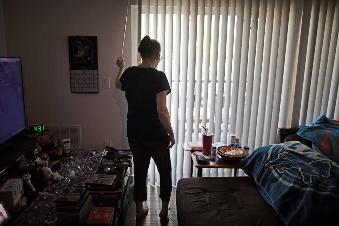 Debbie Willson twists a rod to close long vertical shades in her dimly lit home. Her back is to the camera.