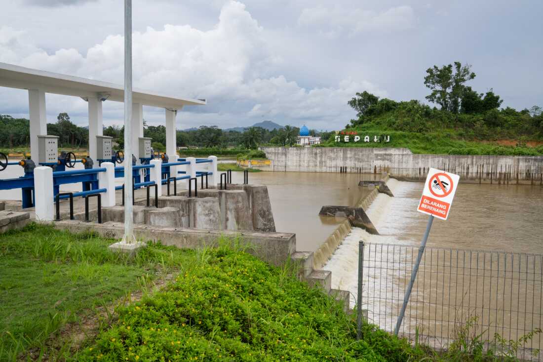 تتدفق المياه عبر سد تم بناؤه حديثًا ومحطة لمعالجة المياه في نوسانتارا. تمتد الأشجار الخضراء وأوراق الشجر عبر الخلفية.