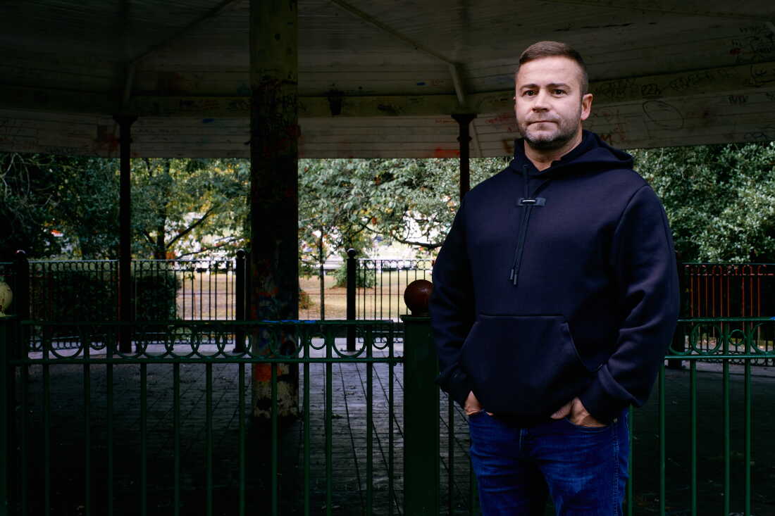 Shane Thomson stands outdoors. He's wearing blue jeans and a dark blue hoodie.