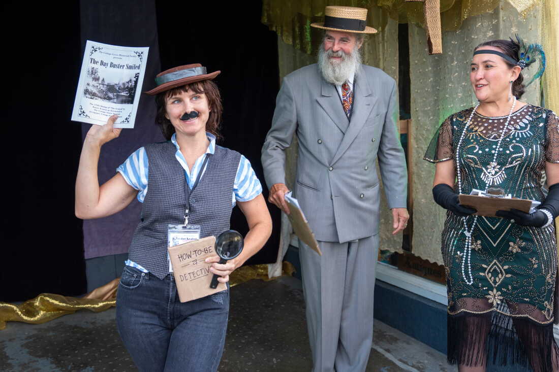 Liz Bartholomew (left) wins for best Buster Keaton look-alike after being awarded by Dana Merryday (center) and Jennifer Ferraez Chartrand during a 1920s themed costume contest. Liz is wearing a porkpie hat, a fake mustache, a vest over a striped short-sleeve button down shirt and carrying magnifying glass and a handwritten sign that says "How to be a detective." Dana is wearing a gray vintage suit and straw hat while Jennifer is wearing a green 1920s-style beaded dress and has a headband with a peacock feather in it.
