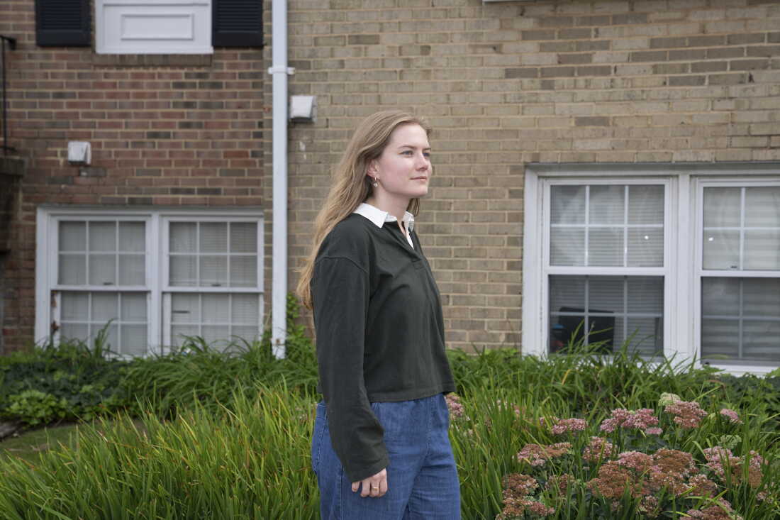 Kasey Hatch stands outdoors. She's wearing blue pants and a black long-sleeved shirt. A multistory brick residential building is in the background.