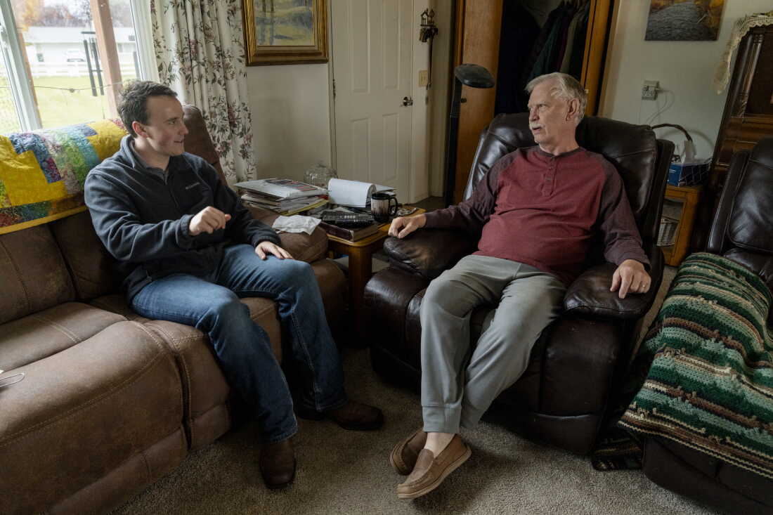 Lukas Schubert (left) speaks with voter Mark Shropshire in Kalispell, Montana. Both men are sitting in Shropshire's living room. Shropshire has gray hair and a gray mustache.