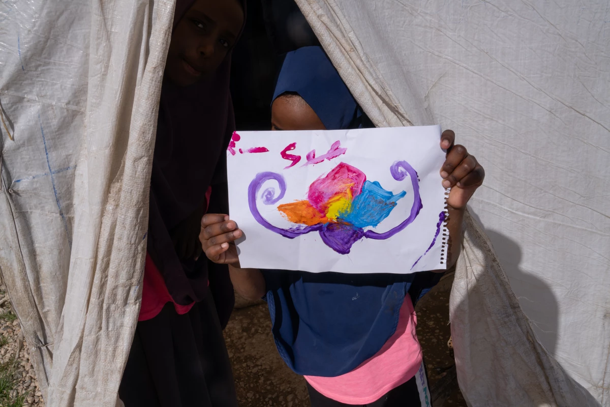 A girl at Roj holds a picture of a flower that she painted. On the back, it says, 'hello friends.'