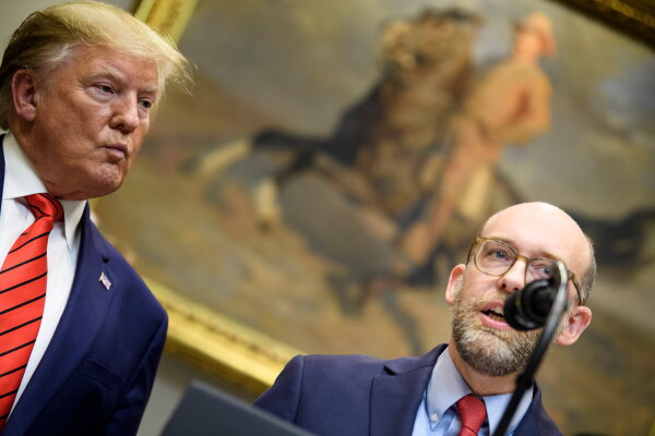 This Oct. 9, 2019, photo shows President Donald Trump on the left and the acting director of the Office of Management and Budget, Russell Vought, on the right. Trump is wearing a blue suit jacket and a red tie with dark stripes. Vought, wearing a blue suit jacket and a red tie, is speaking into a microphone. On the wall behind them is a large painting of a man riding a horse.