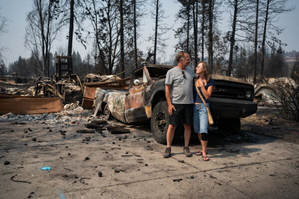 Ken and Shannon Adkins stand in front of their home, which was destroyed by wildfire. A heavily damaged truck sits behind them.